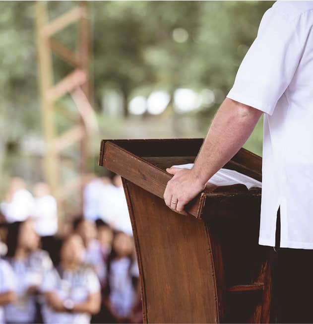 Person in white shirt speaking at outdoor event with audience in background.