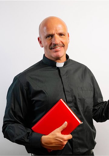 Clergy man in black clerical shirt holding red book, smiling, against plain background.
