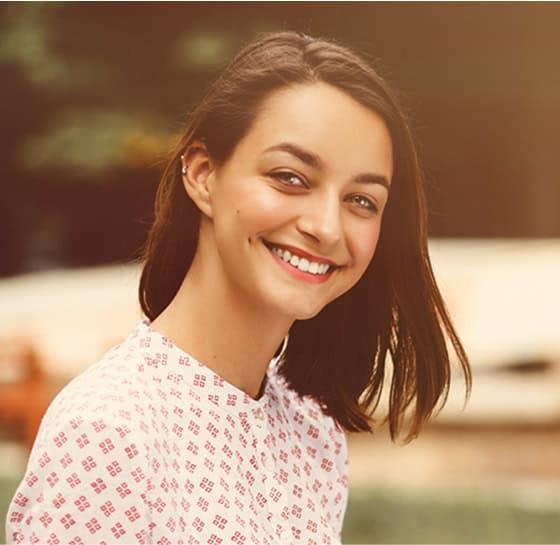 Smiling woman with brown hair in a pink patterned top outdoors.