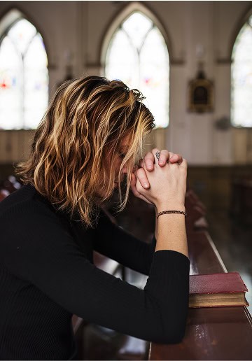 Woman praying in church with hands clasped and head bowed, serene atmosphere.